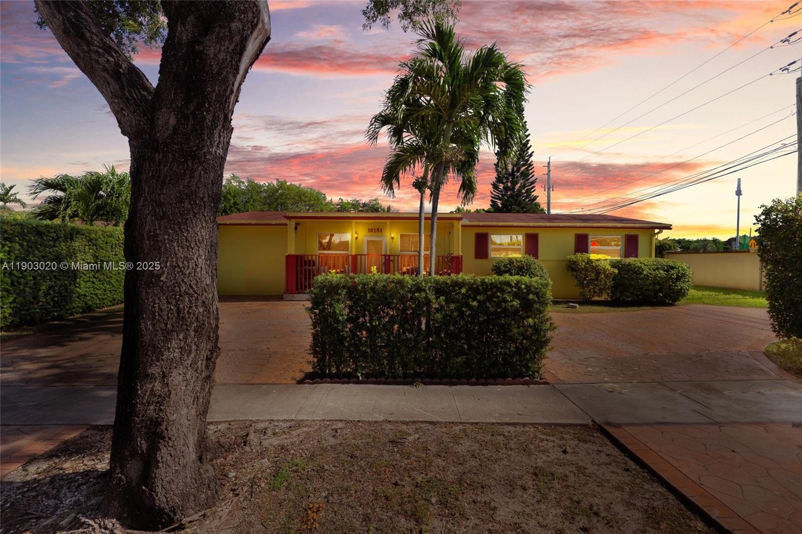 A yellow single-story house with a paved driveway and a hedge sits under a sunset — 18151 NE 9th Ct, North Miami Beach FL 33162, NORTH MIAMI BEACH, FL