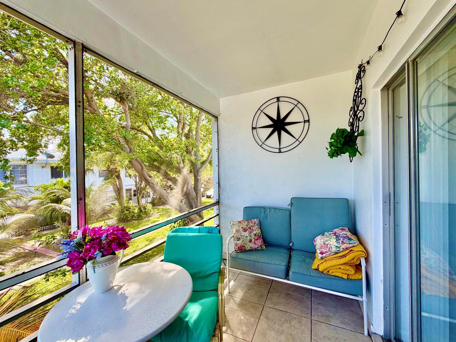 A screened-in balcony features a teal loveseat, a small round table, and a wall-mounted compass — 2551 W Golf Boulevard # 204, Pompano Beach FL 33064, POMPANO BEACH, FL