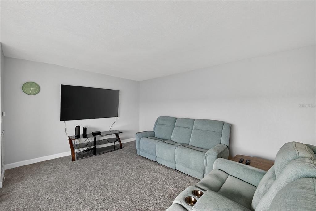 A living room features gray carpeting, light gray walls, a wall-mounted television, and two blue sofas — 5521 80TH STREET N UNIT 511, ST PETERSBURG, FL