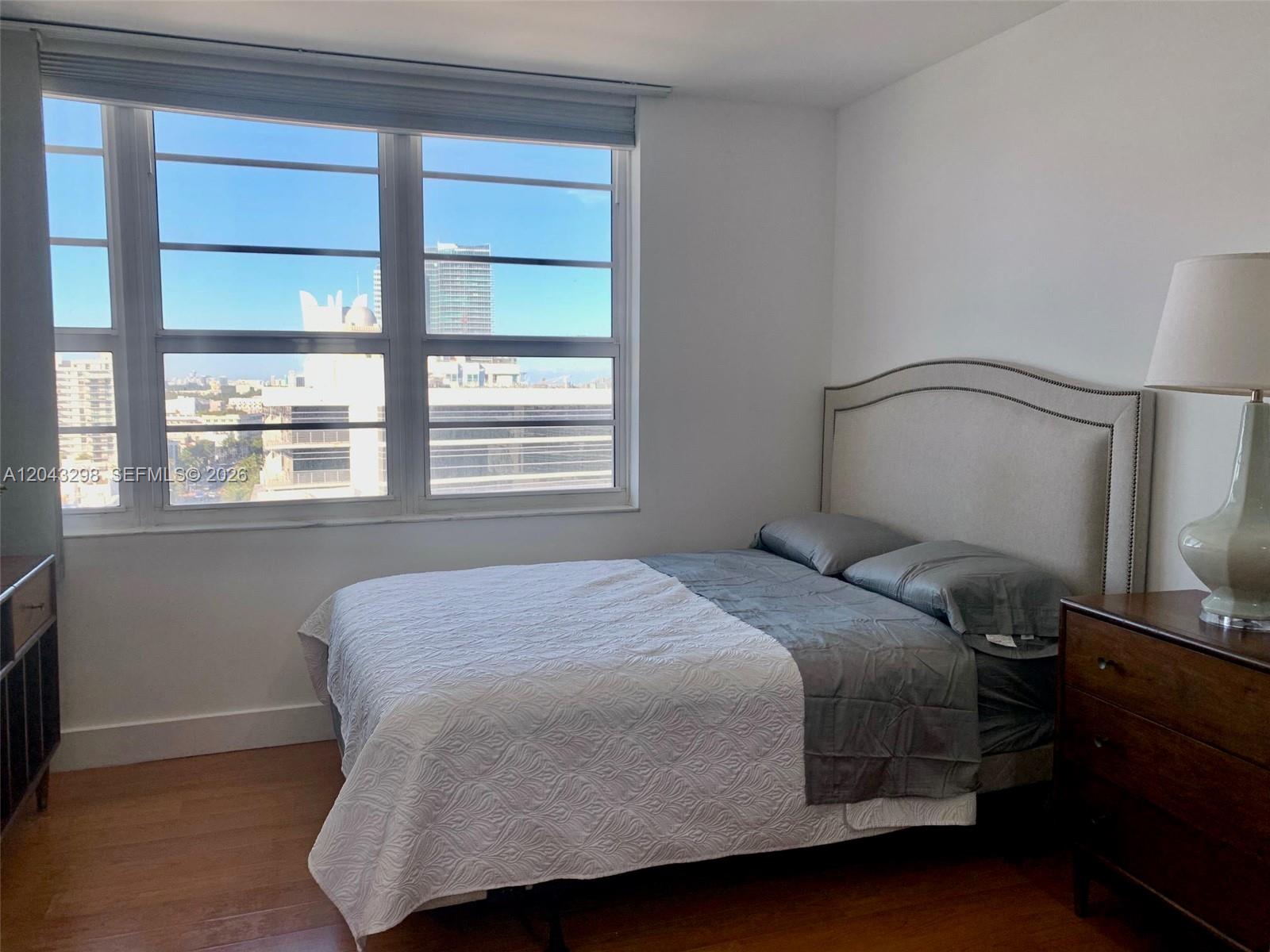 A bedroom features a bed with a grey headboard, wood flooring, and a large window — 100 Lincoln Rd # 1514A, Miami Beach FL 33139, MIAMI BEACH, FL
