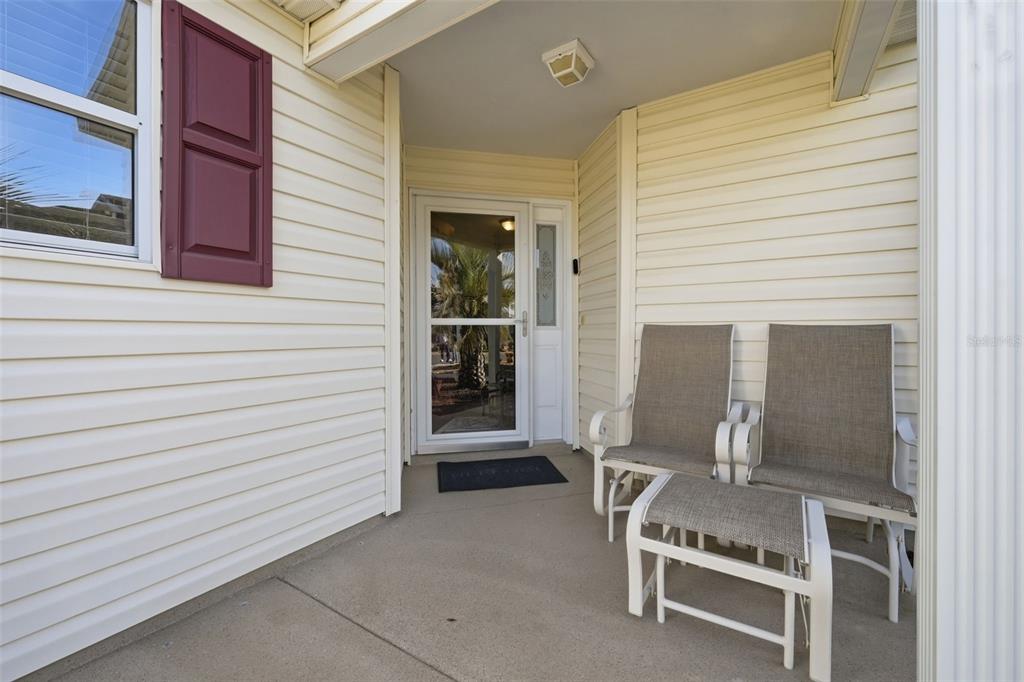 A covered front porch features beige siding, a glass storm door, and two outdoor chairs — 2311 CRESTVIEW STREET, THE VILLAGES, FL