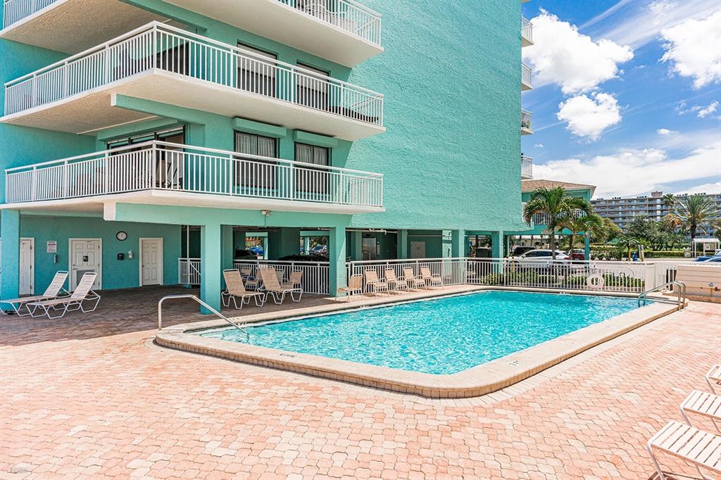 A turquoise multi-story building features balconies overlooking a rectangular swimming pool on a paved deck — 19440 GULF BOULEVARD UNIT 105, INDIAN SHORES, FL