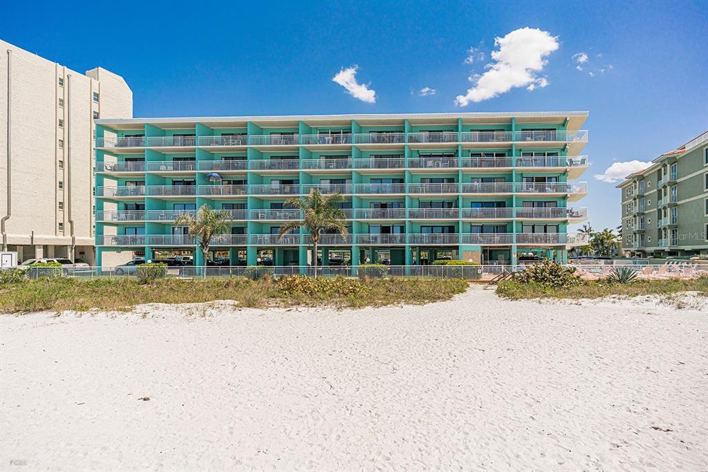 A multi-story turquoise apartment building stands behind a sandy beach under a clear blue sky — 19440 GULF BOULEVARD UNIT 105, INDIAN SHORES, FL