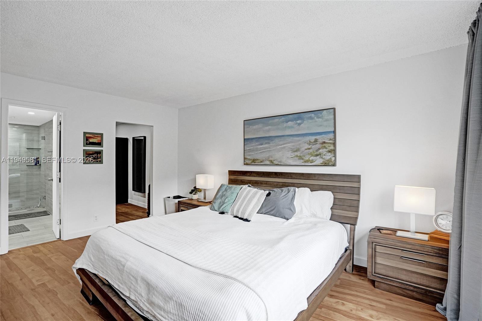 A bedroom features a wooden bed frame, white bedding, two nightstands, and an attached bathroom — 401 Golden Isles Dr # 302, Hallandale Beach FL 33009, HALLANDALE BEACH, FL