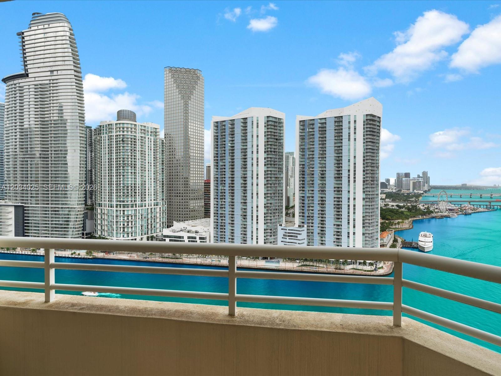 A balcony overlooks a waterfront cityscape featuring several high-rise buildings and a bright blue bay — 888 Brickell Key Dr # 2510, Miami FL 33131, MIAMI, FL