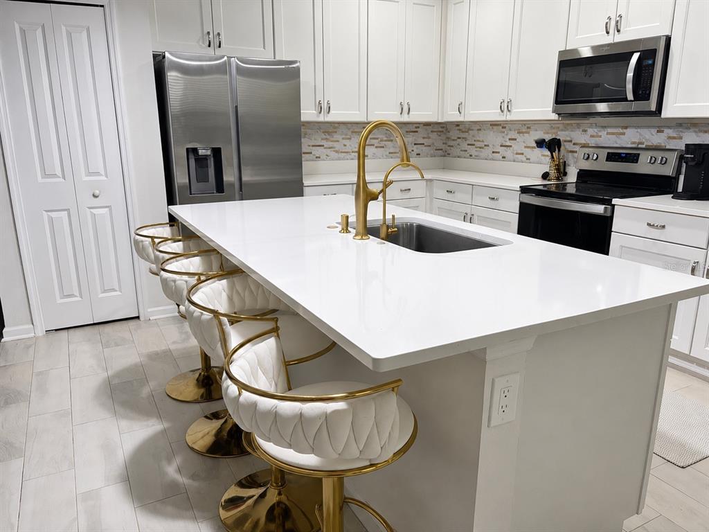 A modern kitchen features white cabinetry, a large quartz island with gold stools, and stainless appliances — 10972 BRIGHTLEAF COURT, RIVERVIEW, FL