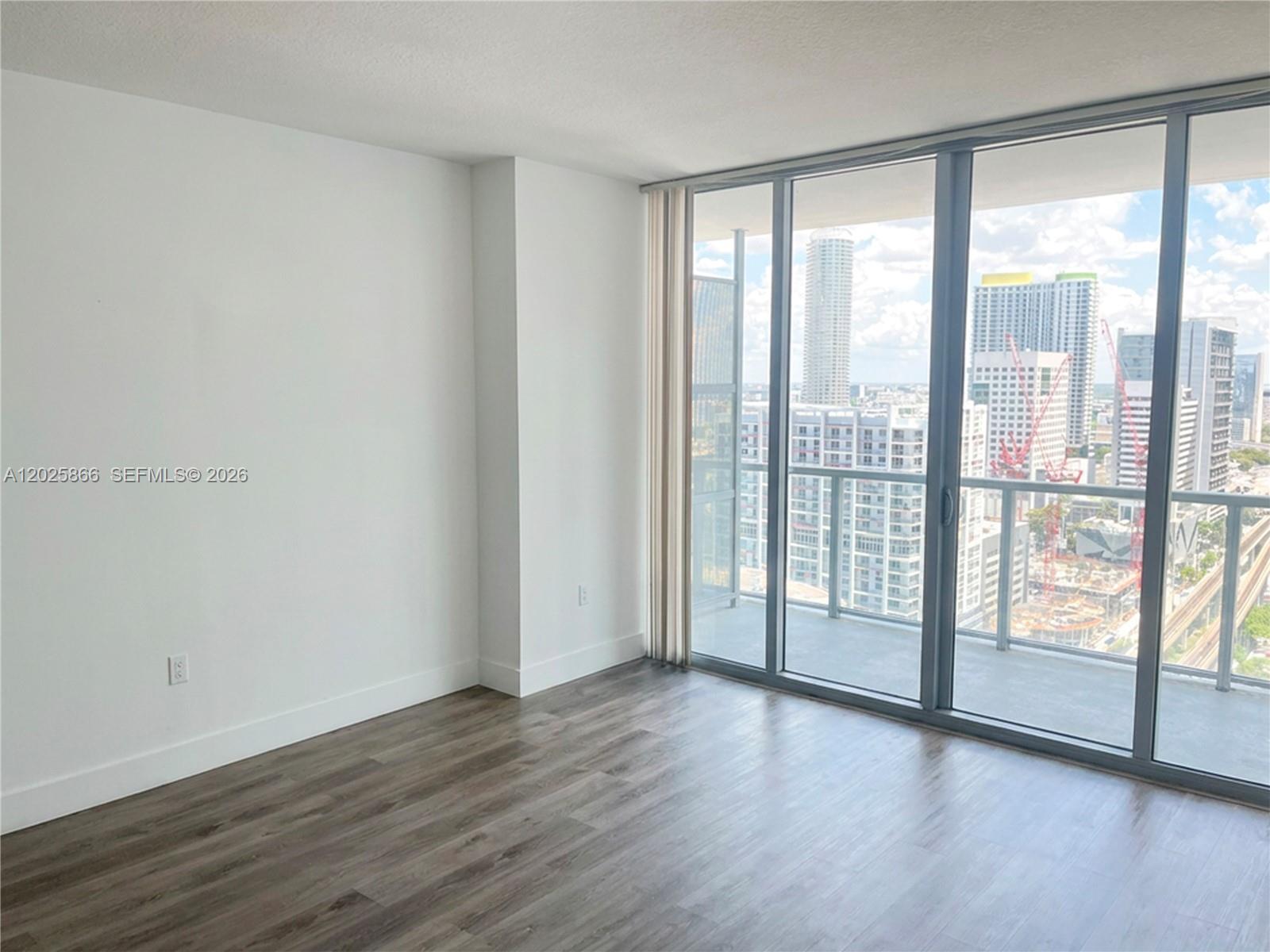 An empty room features wood-look flooring, white walls, and floor-to-ceiling windows overlooking a city skyline — 79 SW 12 ST, Miami FL 33130, MIAMI, FL