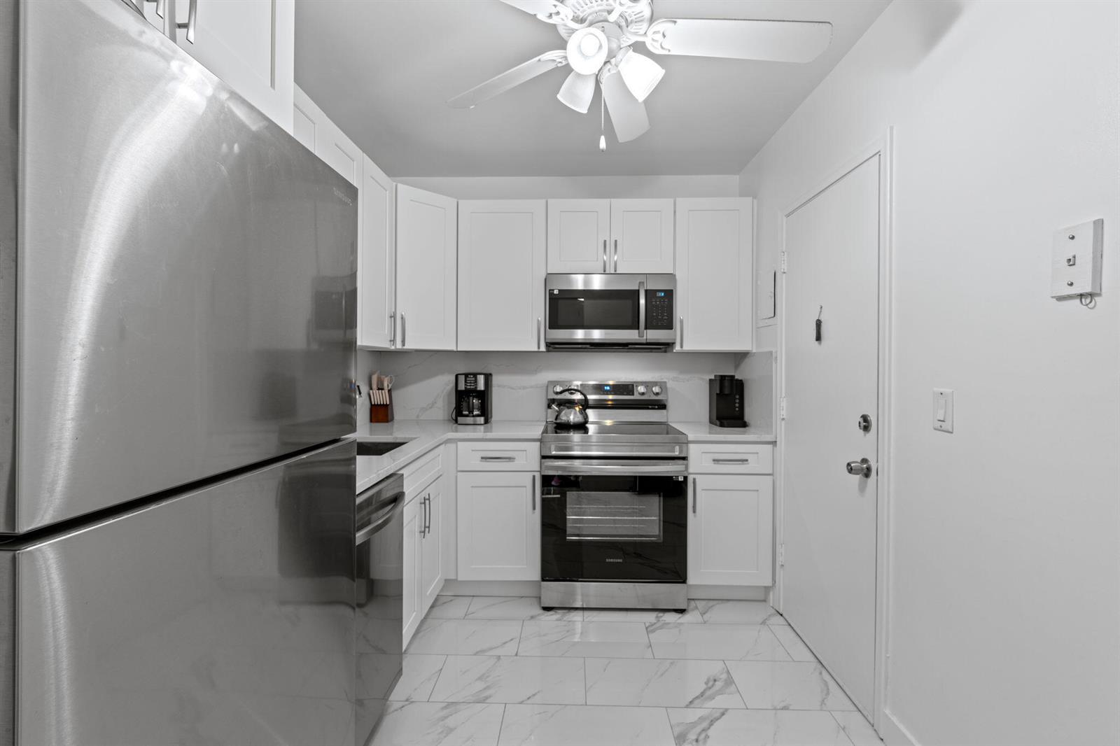 A small kitchen features white cabinets, stainless steel appliances, and white marble-patterned tile flooring — 418 S Cypress Road # 128, Pompano Beach FL 33060, POMPANO BEACH, FL