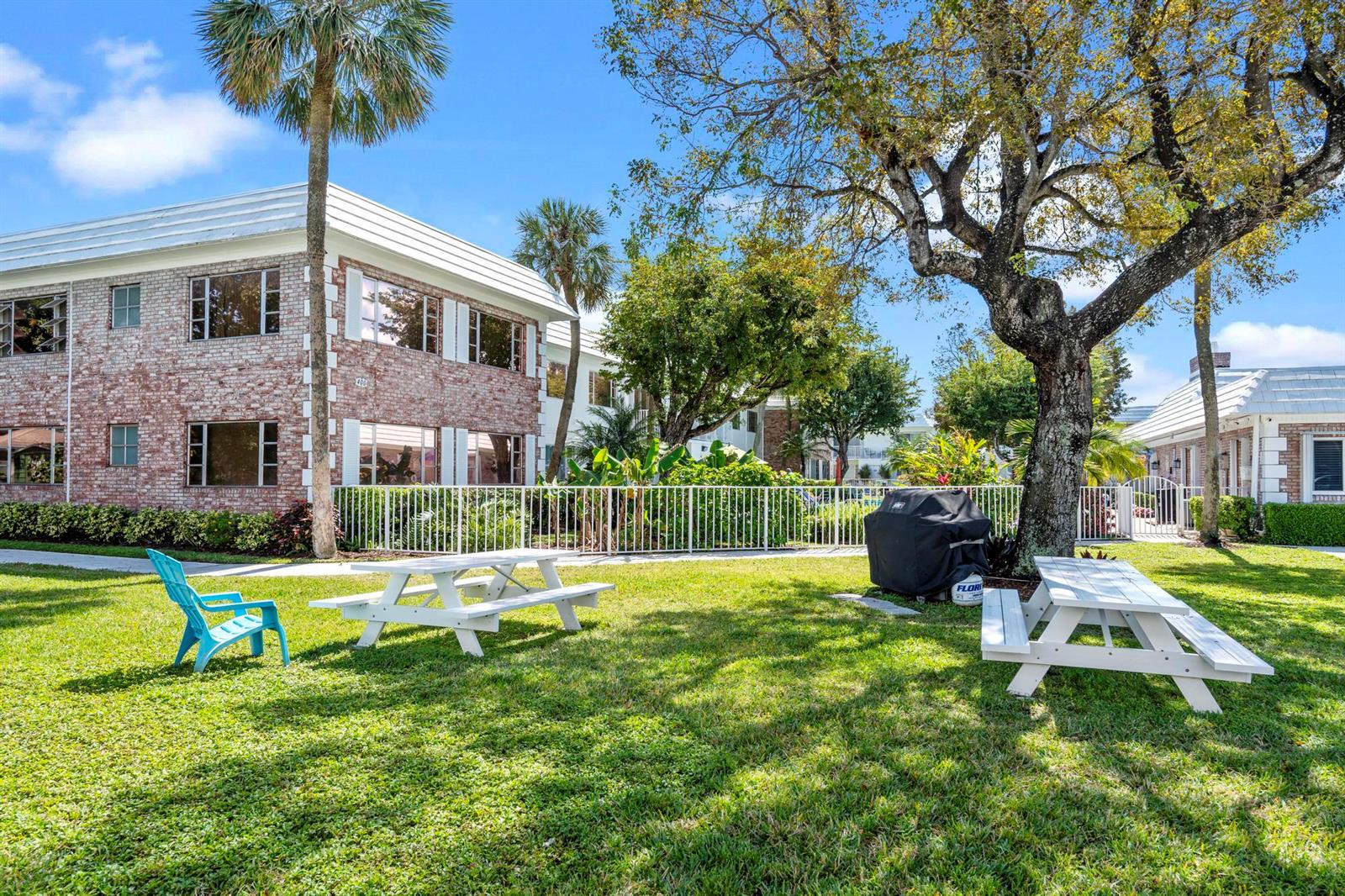 A grassy courtyard features two white picnic tables, a blue chair, and a brick building — 418 S Cypress Road # 128, Pompano Beach FL 33060, POMPANO BEACH, FL
