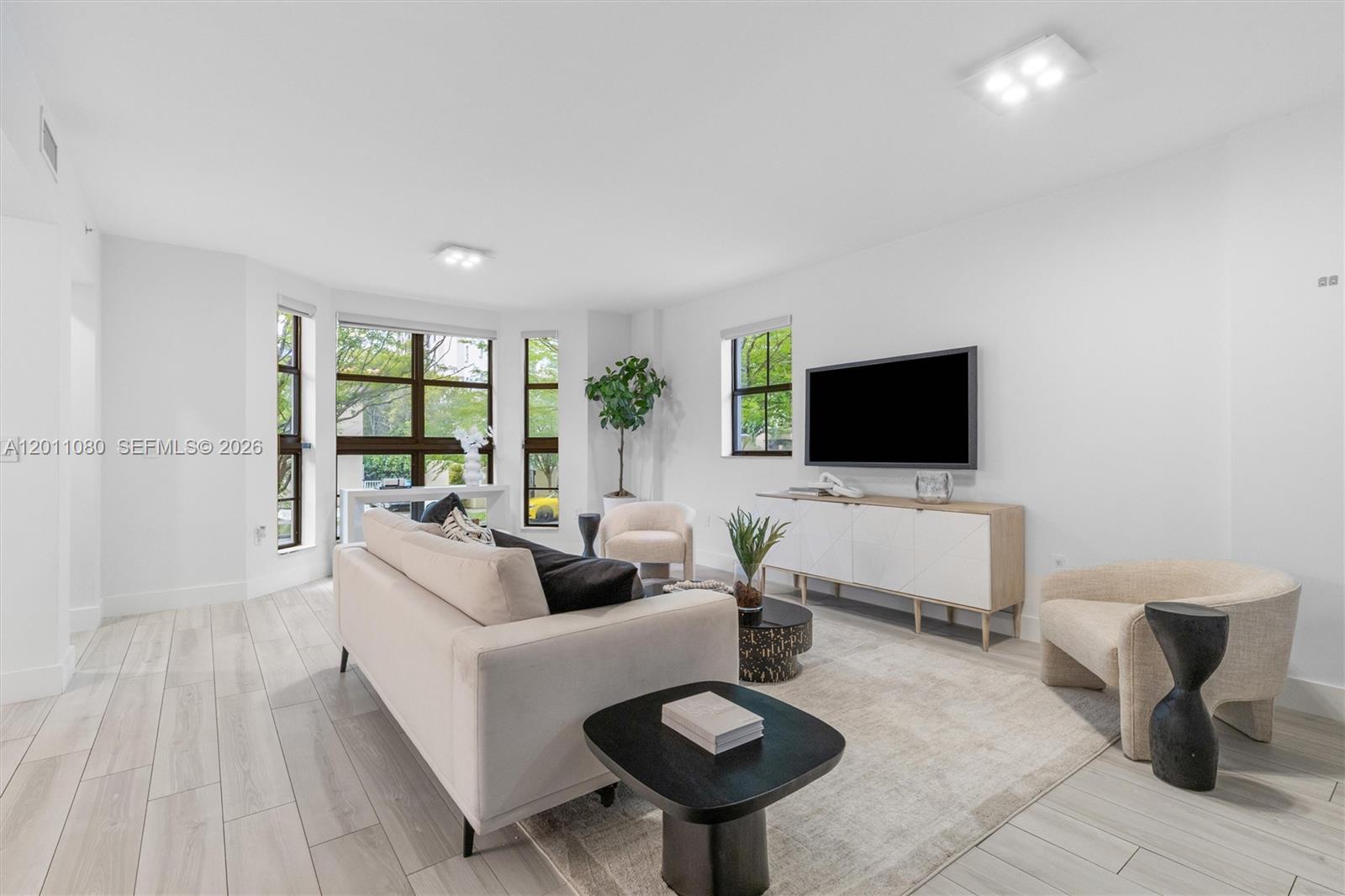 A bright living room features light wood-look flooring, a white sofa, and a wall-mounted television — 1650 Galiano St, Coral Gables FL 33134, CORAL GABLES, FL