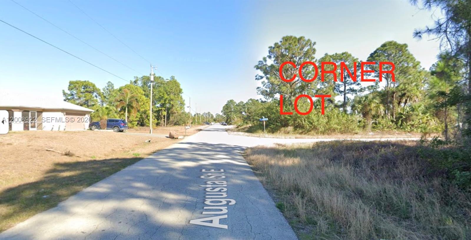 A paved road leads toward a vacant, wooded corner lot under a clear blue sky — 507 POWELL AVE S, Lehigh Acres FL 33974, LEHIGH ACRES, FL