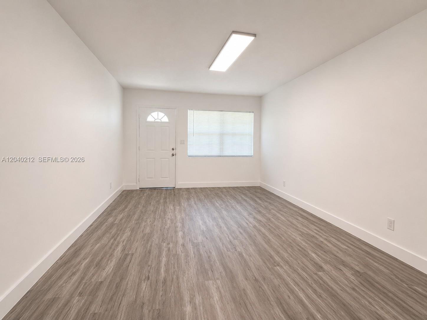 An empty room features white walls, gray wood-look flooring, a window, and a door — 730 SW 5th St # 1, Hallandale Beach FL 33009, HALLANDALE BEACH, FL