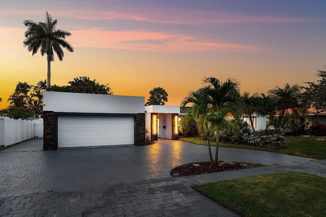 A modern house exterior with a stone-accented entryway, paved driveway, and palm trees at sunset — 2611 NE 49 Street, Lighthouse Point FL 33064, LIGHTHOUSE POINT, FL