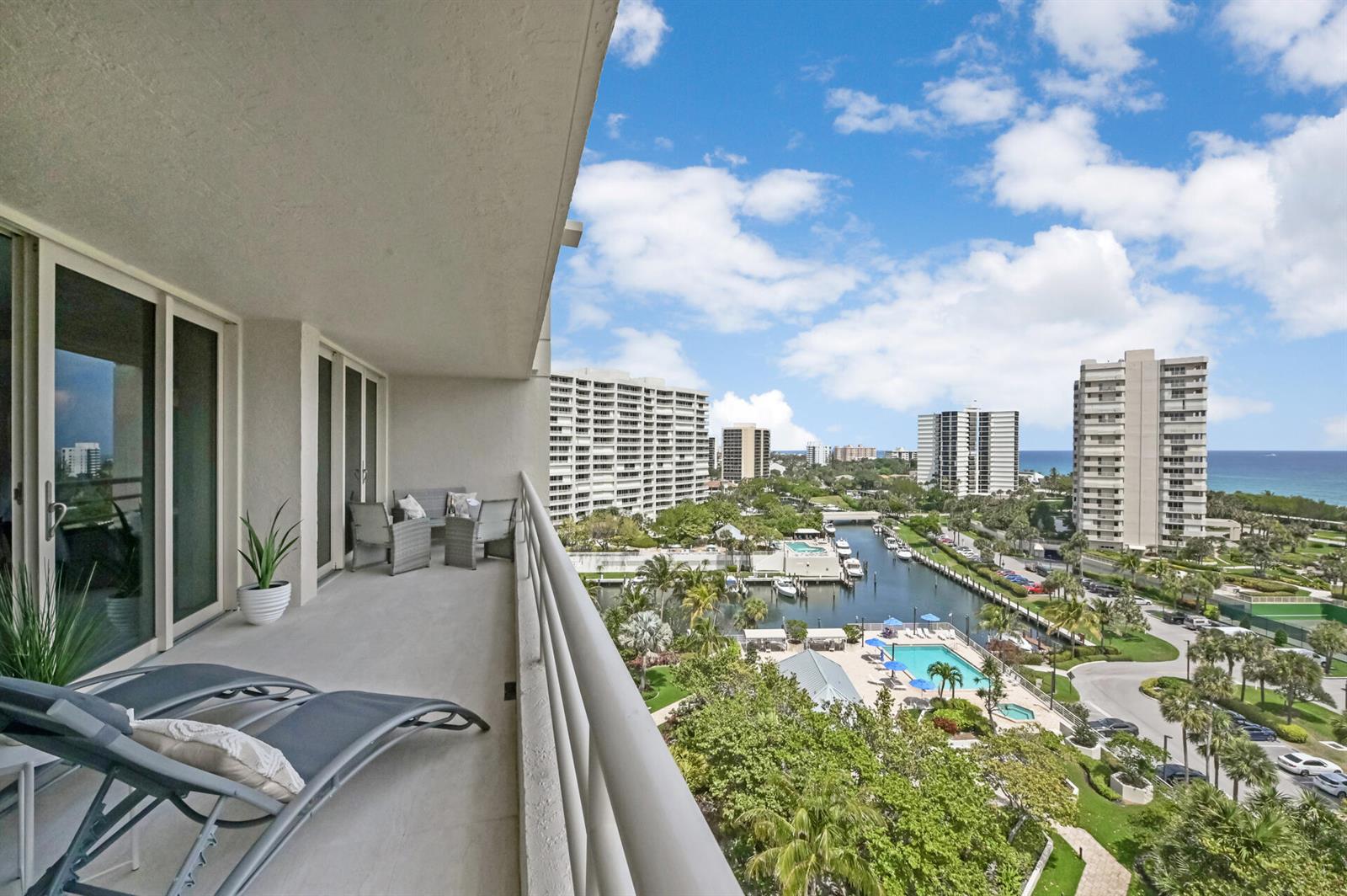 A balcony with lounge chairs overlooks a canal, swimming pool, and high-rise buildings near the ocean — 4101 N Ocean Boulevard Unit 1008, Boca Raton FL 33431, BOCA RATON, FL