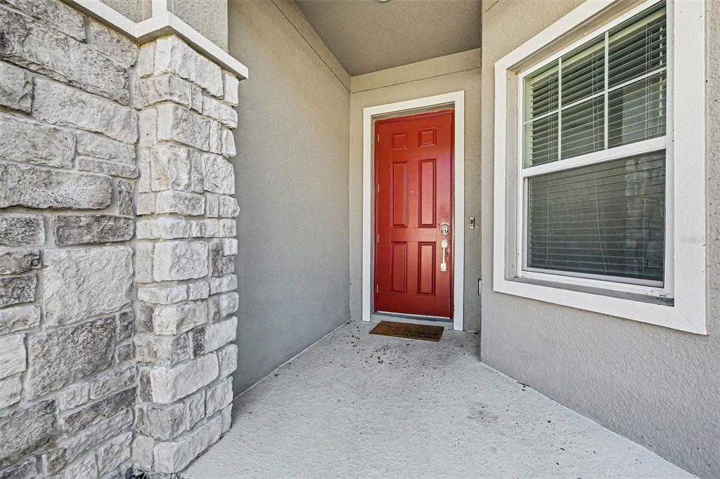 A red front door is set within a covered porch featuring stone pillar accents — 6079 CLAYSTONE WAY, MOUNT DORA, FL
