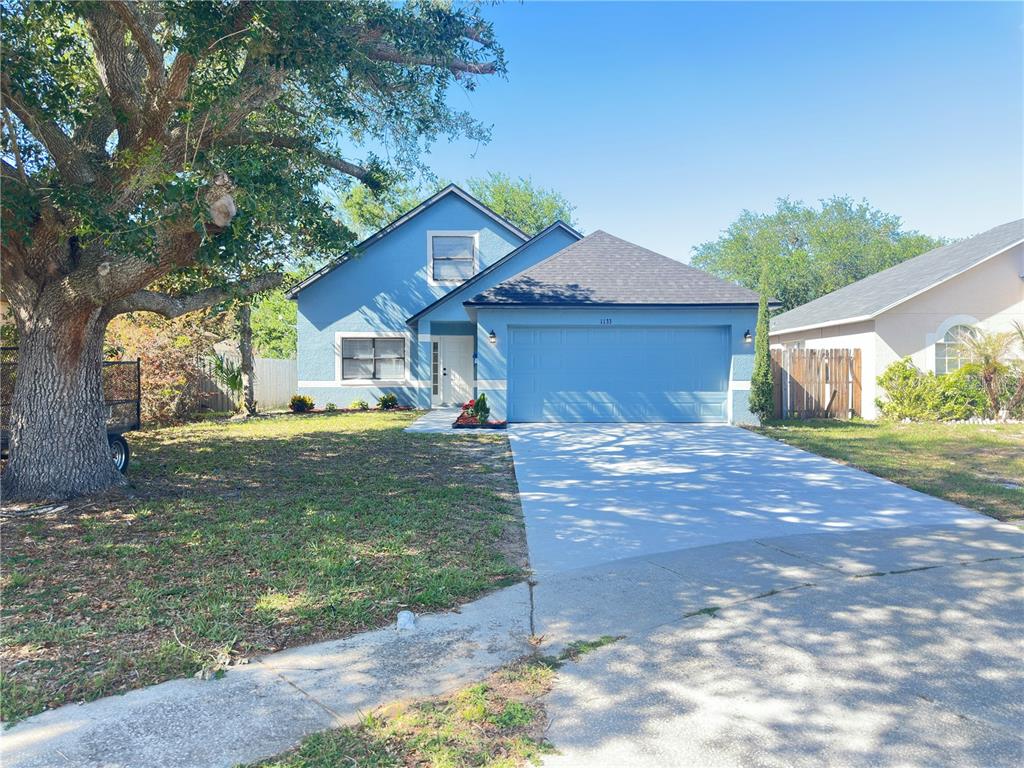 A blue two-story house features a front-facing garage, a concrete driveway, and a large tree — 1133 OLYMPIC COURT, APOPKA, FL