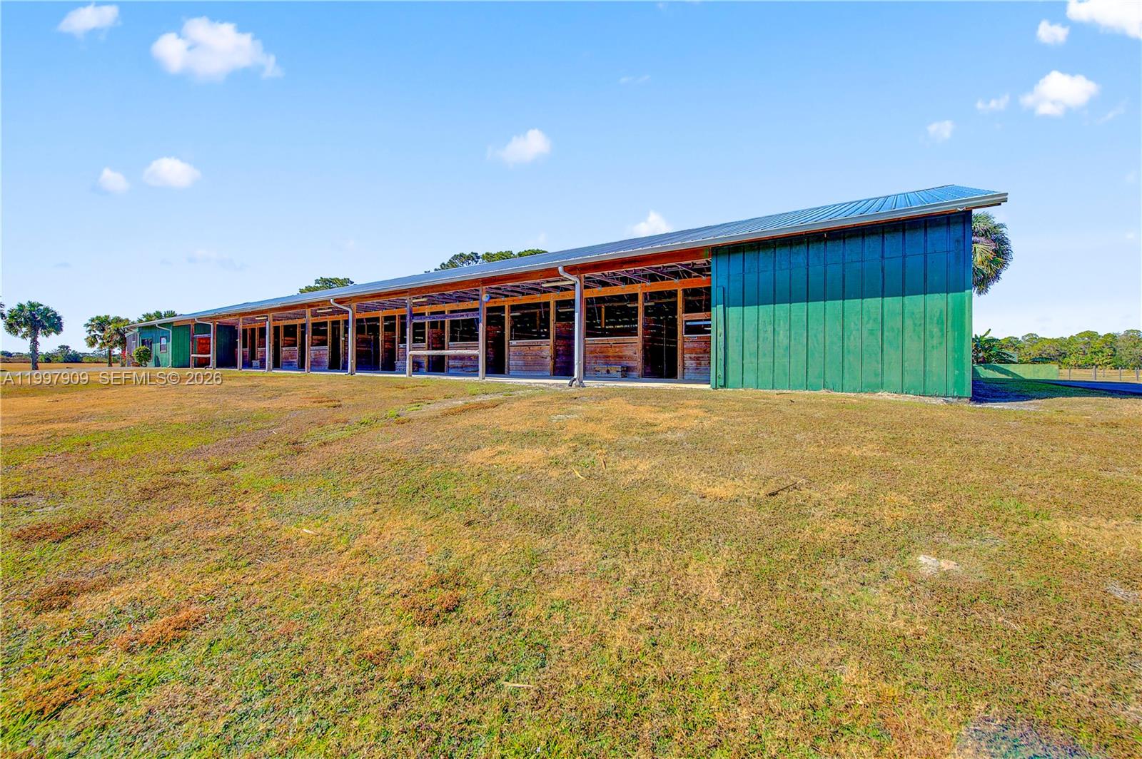A long, green-sided barn with multiple open horse stalls sits on a grassy field — 30003 SW Martin Hwy, Okeechobee FL 34974, OKEECHOBEE, FL
