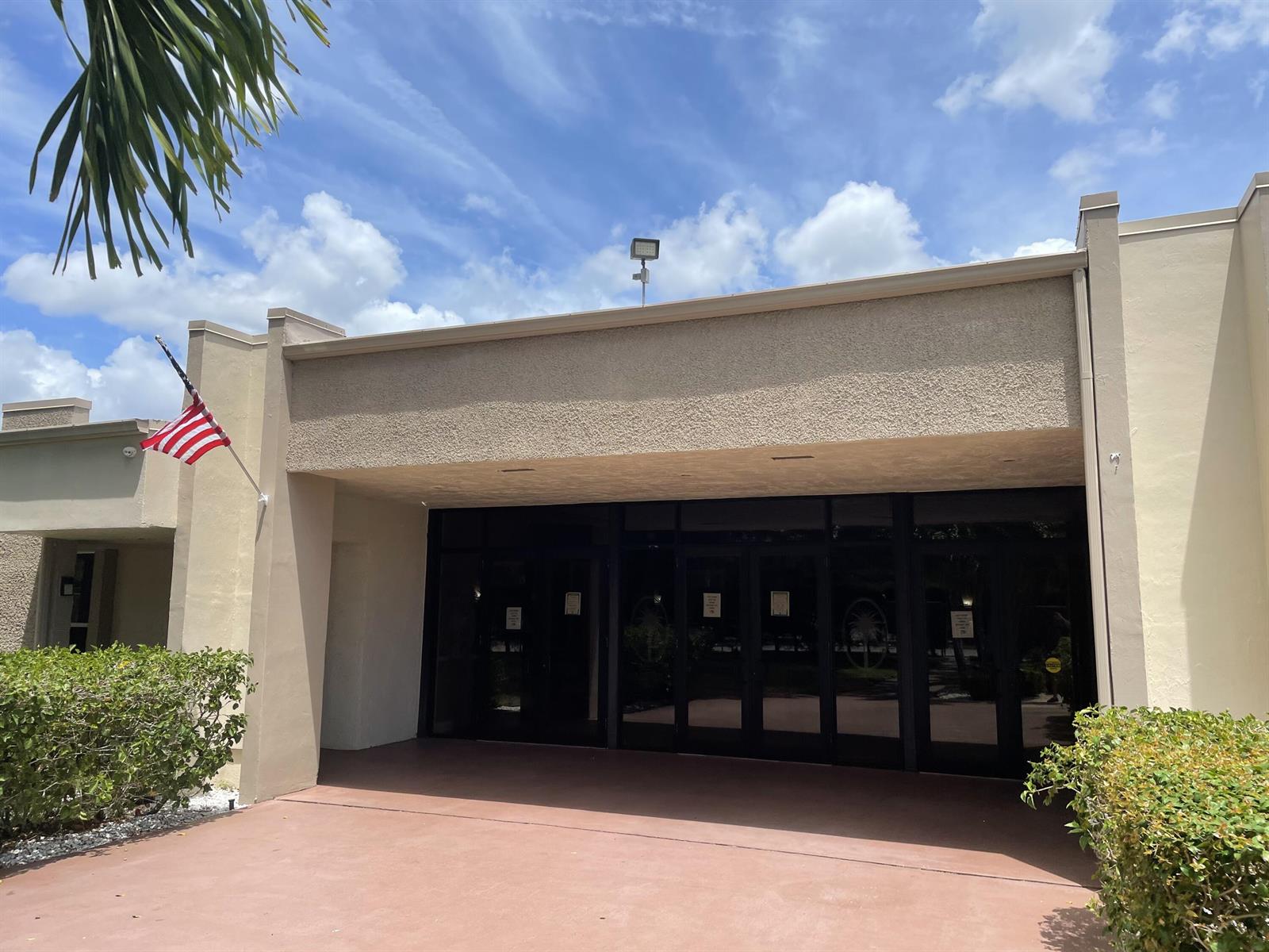 The exterior of a beige building features a covered entrance with glass doors and a flag — 705 Lori Drive # 315, Palm Springs FL 33461, PALM SPRINGS, FL