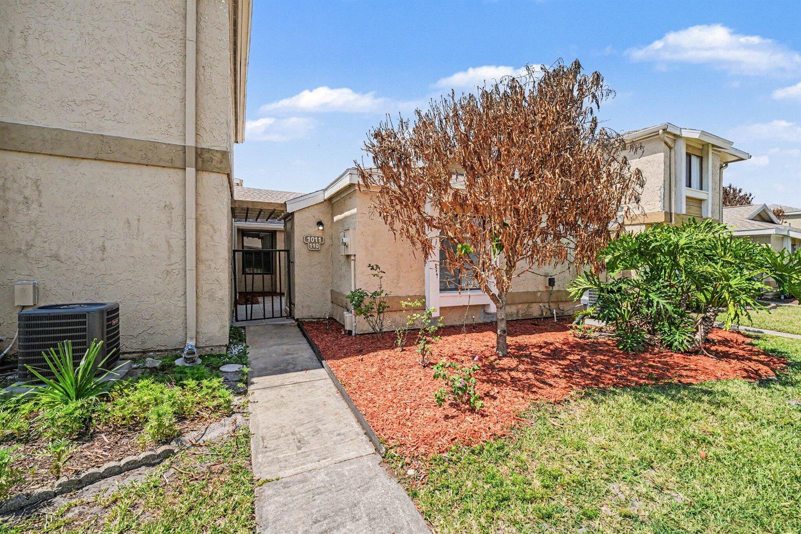 A beige stucco house exterior features a concrete walkway, a dead tree, and red mulch — 1011 NE Abada Ct Court NE # 110, Palm Bay FL 32905, PALM BAY, FL