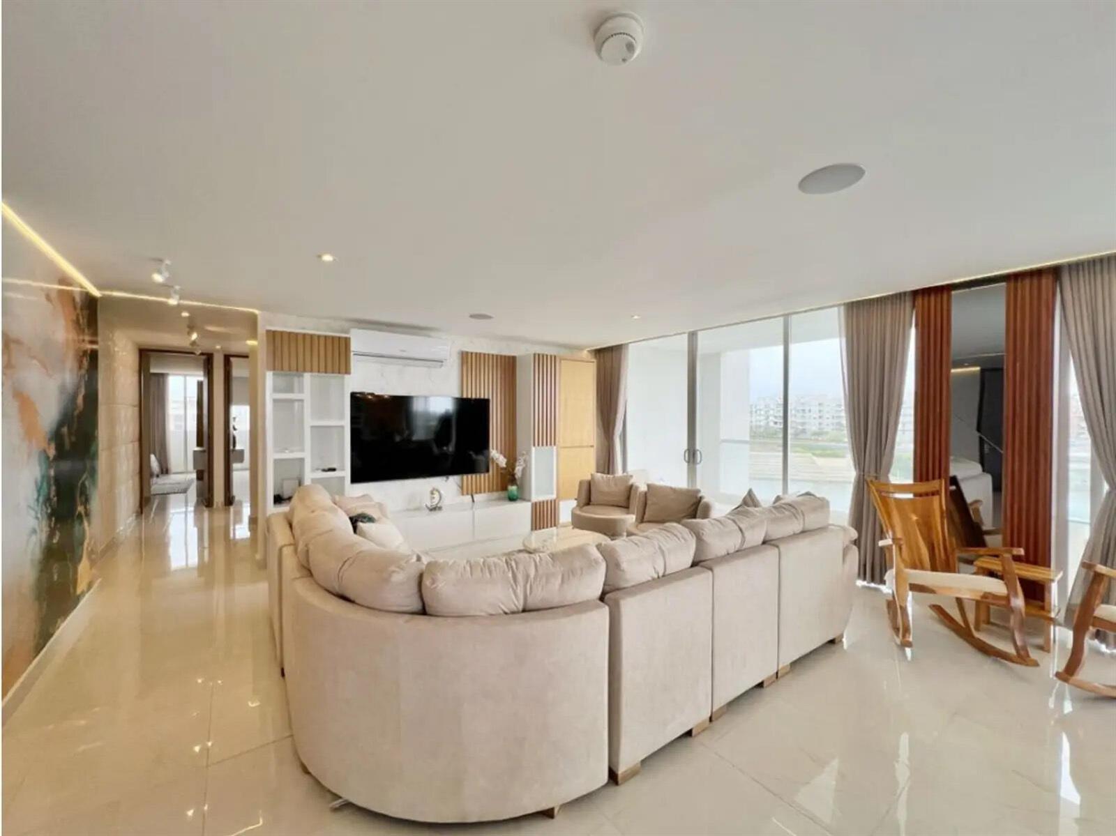 A living room features a large beige sectional sofa, a wall-mounted television, and floor-to-ceiling windows — Km 8 Via Al Mar, FL
