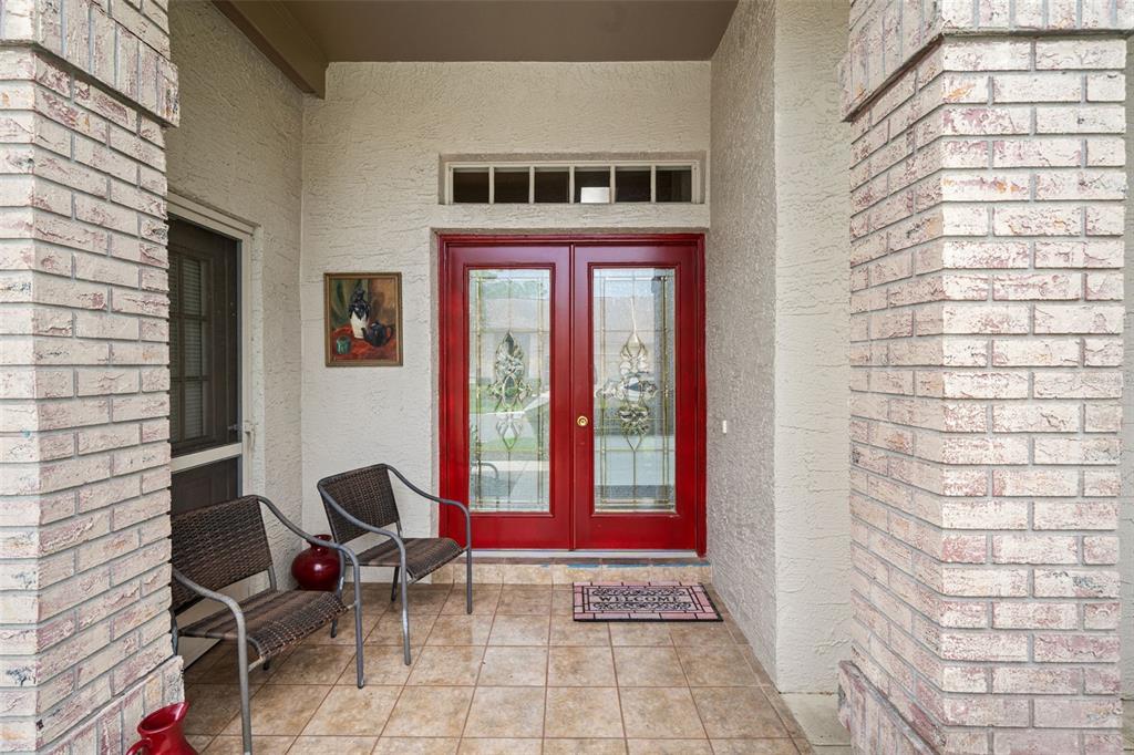 A covered front porch features tiled flooring, two wicker chairs, and a red double front door — 7392 MEADOW BROOK LANE, SPRING HILL, FL