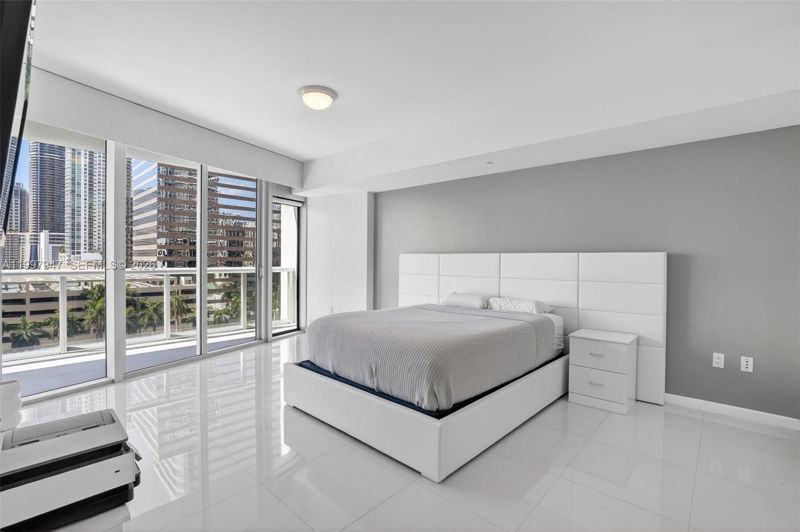 A modern bedroom features a white bed, gray accent wall, and floor-to-ceiling windows with city views — 495 Brickell Ave # 1007, Miami FL 33131, MIAMI, FL