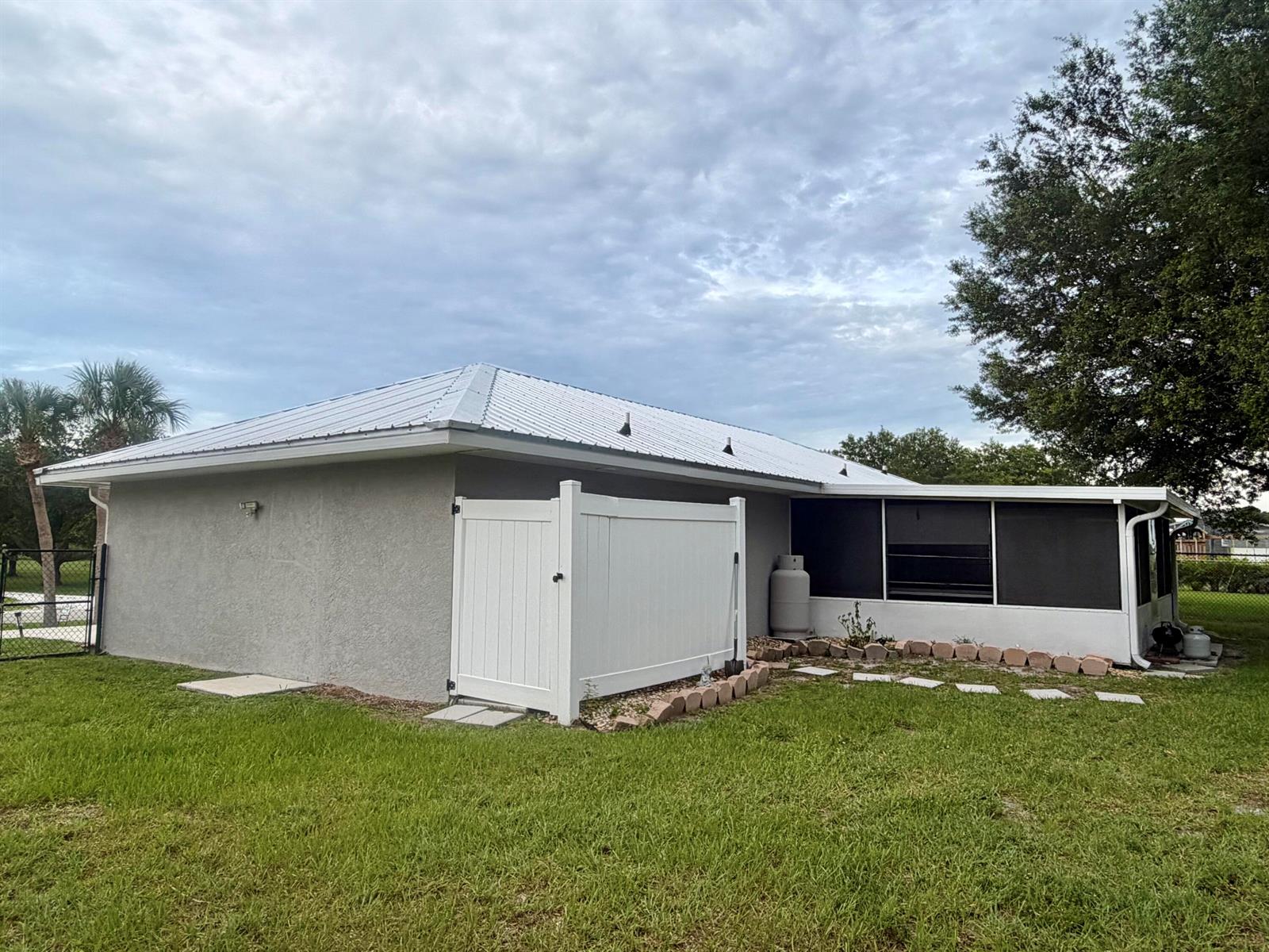 A gray stucco house features a white metal roof, a screened porch, and a privacy fence — 7823 SW 9th Street, Okeechobee FL 34974, OKEECHOBEE, FL