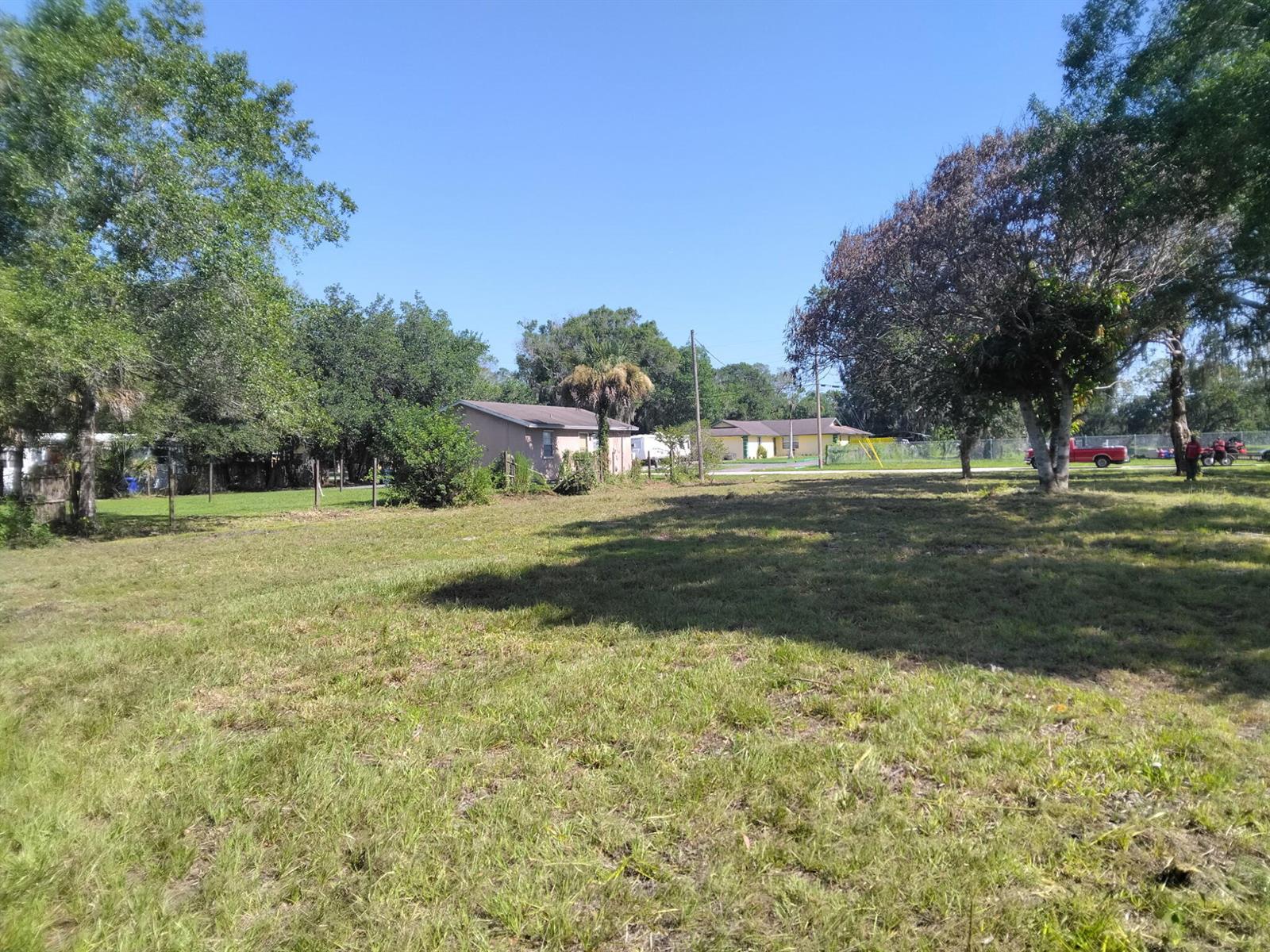 A grassy, open lot features scattered trees and neighboring houses under a clear blue sky — FELLSMERE, FL