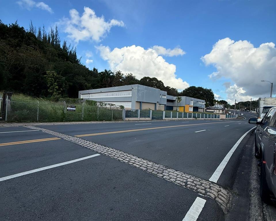A gray commercial building sits behind a chain-link fence along a paved road — AVE ORQUIDEA URB VALENCIA, BAYAMON, PR