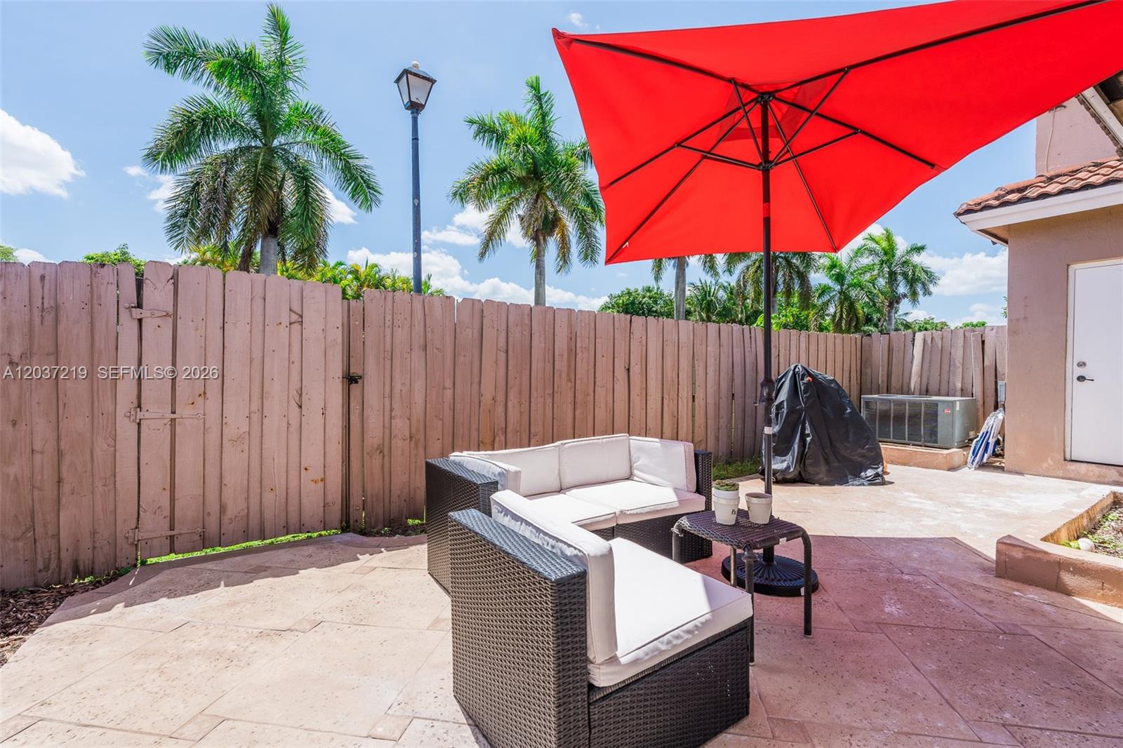 A patio features a red umbrella, wicker furniture, and a wooden fence under palm trees — 10297 SW 162nd Ct, Miami FL 33196, MIAMI, FL