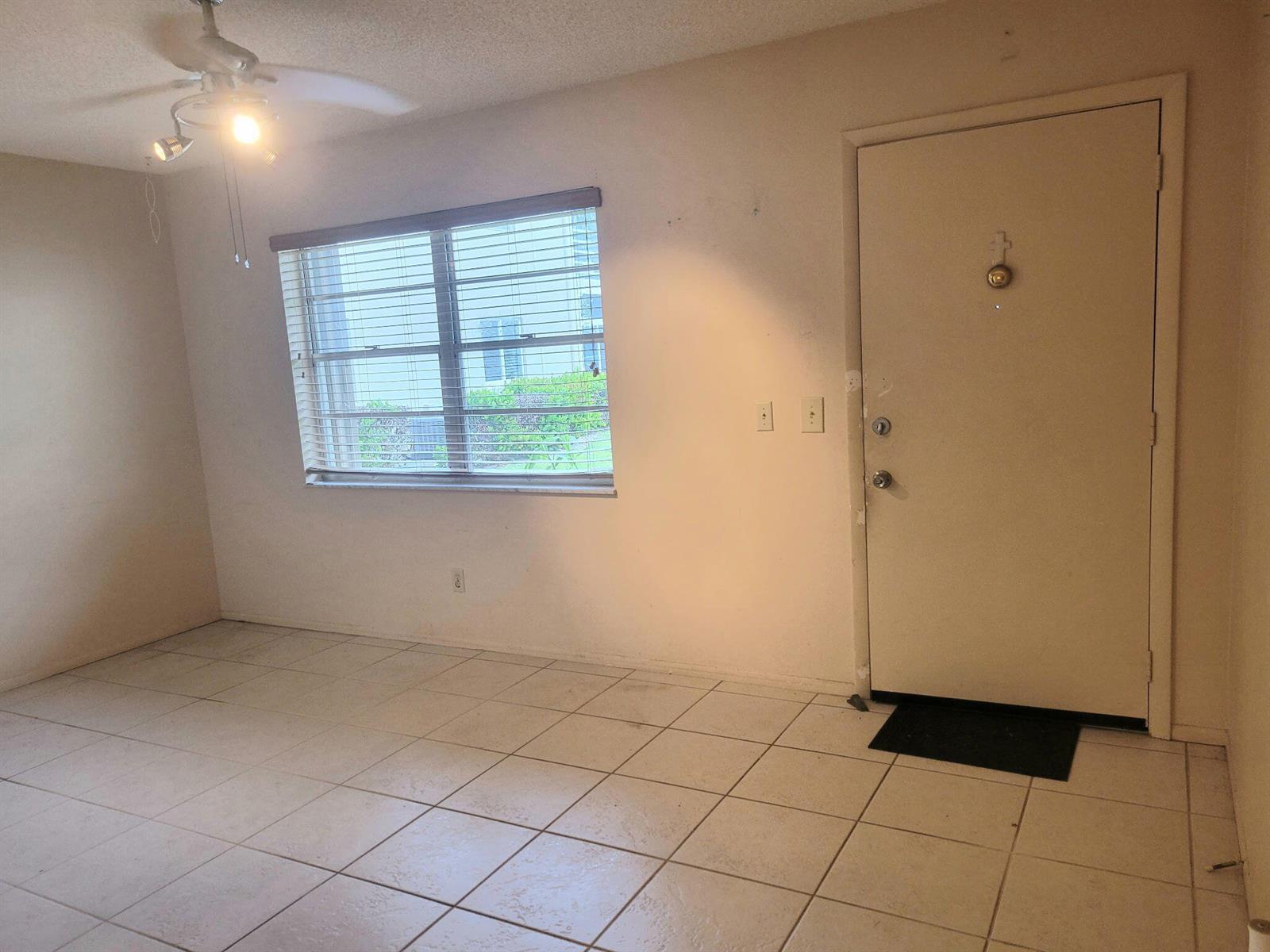 An empty room with beige tile flooring, a ceiling fan, a window, and an entry door — 26 Lake Vista Trail # 104, FL 34952, FL