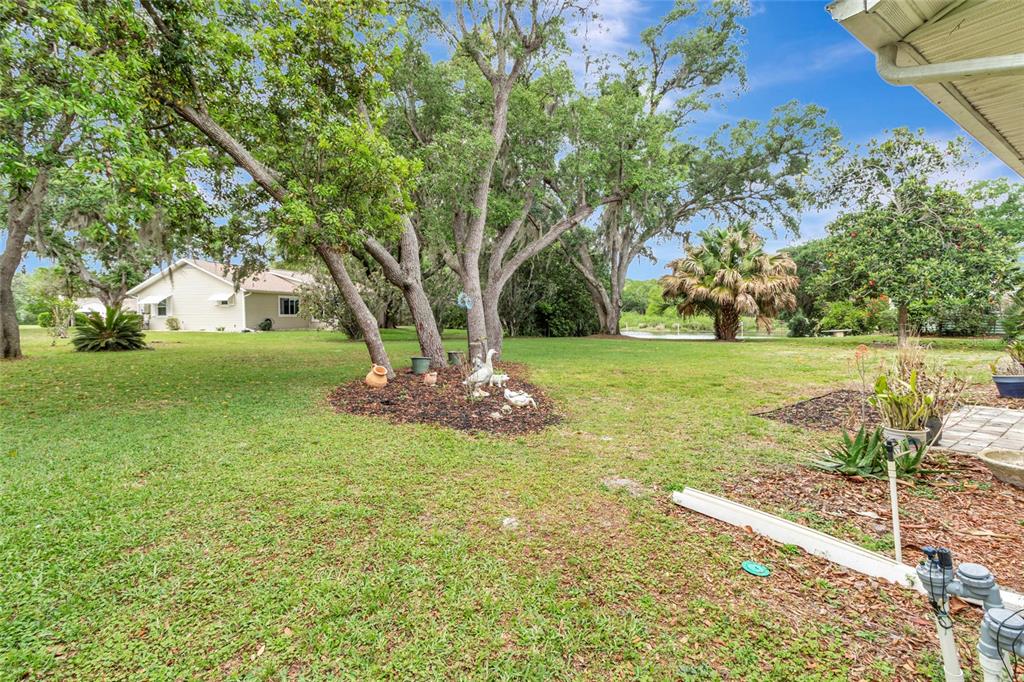 A grassy backyard features large trees, a mulch bed with garden ornaments, and a pond — 27 GORDON COURT, LEESBURG, FL