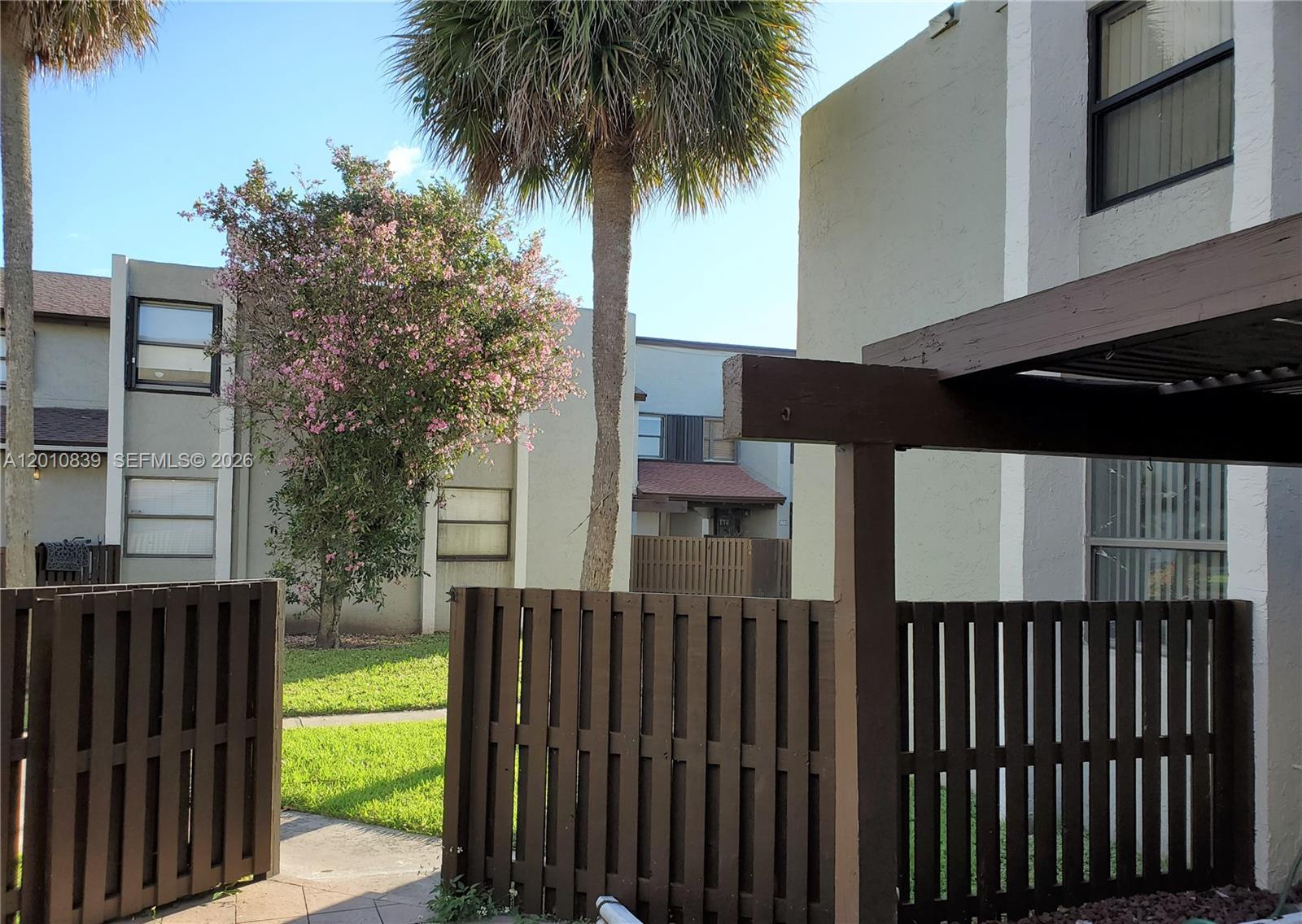 A brown wooden fence surrounds a patio area with a palm tree and flowering tree — 10020 NW 9th St Cir # 102, Miami FL 33172, MIAMI, FL