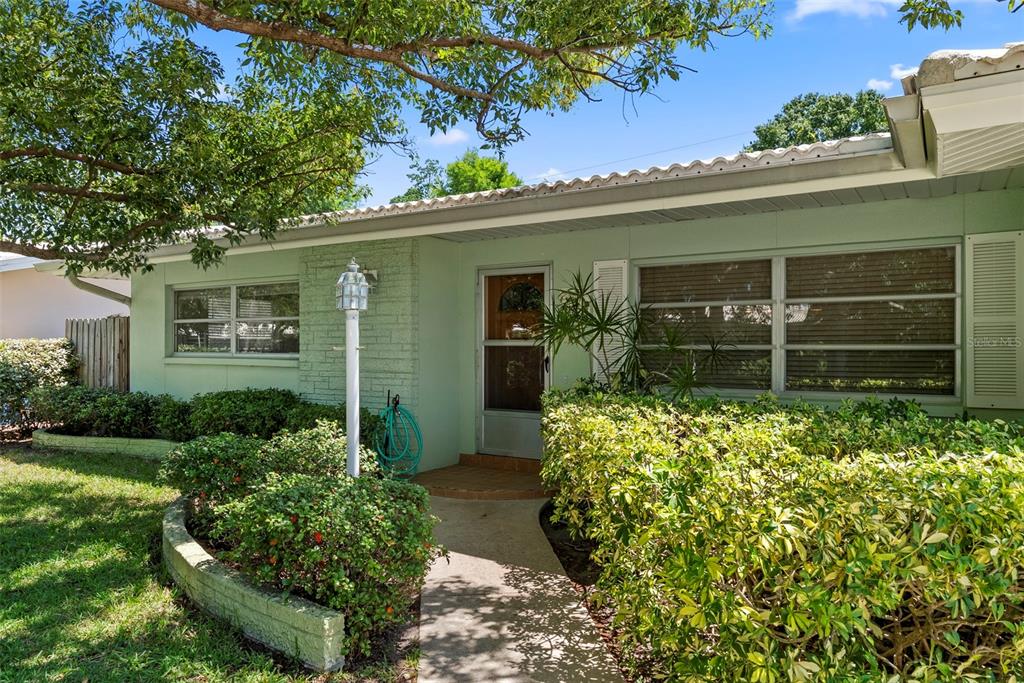 A light green single-story house features a brick accent wall, a walkway, and landscaped bushes — 1413 EMBASSY DRIVE, CLEARWATER, FL