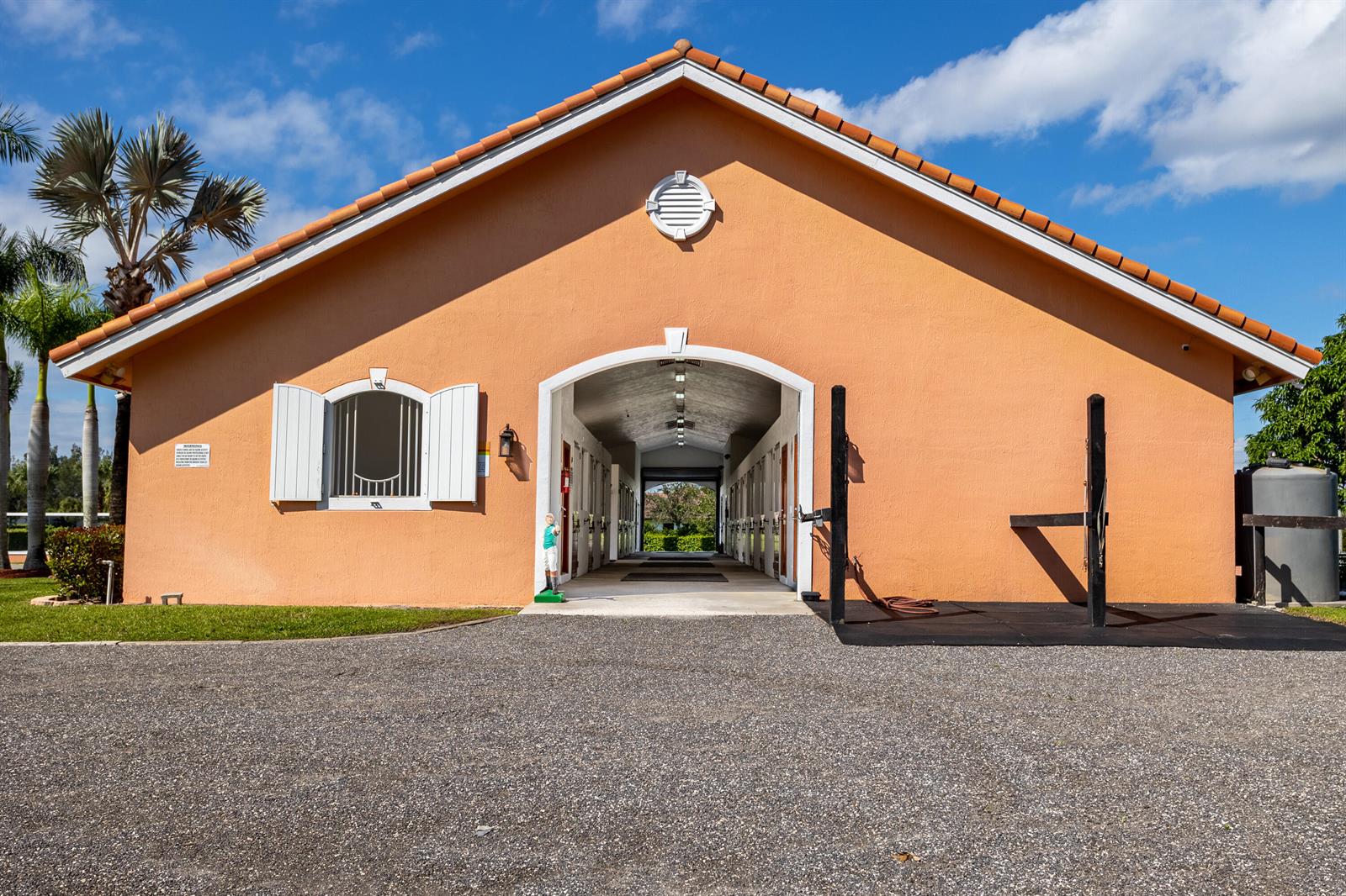 An exterior view of a peach-colored horse stable with a central breezeway and gravel driveway — 4750 South Road, Wellington FL 33414, WELLINGTON, FL