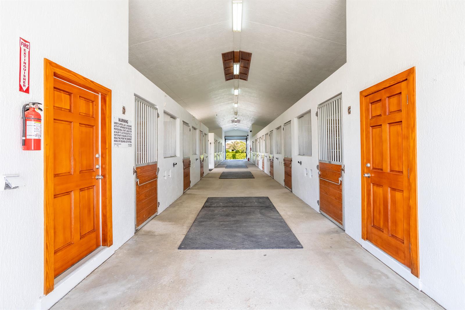 A long, central aisle of a horse stable features wooden doors and metal stall gates — 4750 South Road, Wellington FL 33414, WELLINGTON, FL