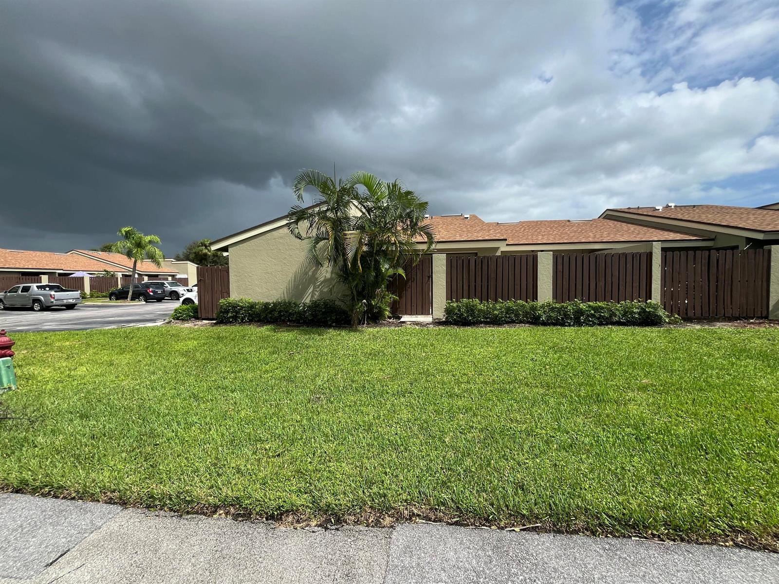 A grassy lawn sits in front of a tan building with a brown fence — 172 Meadows Drive, Boynton Beach FL 33436, BOYNTON BEACH, FL