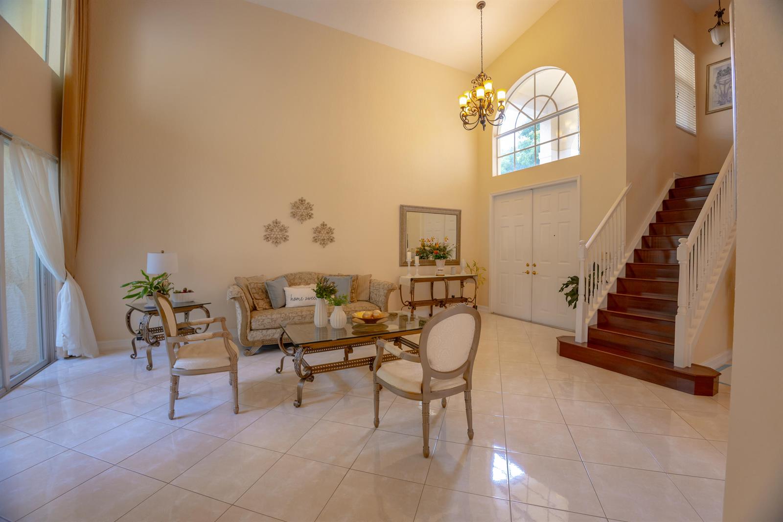 A bright living room features high ceilings, a chandelier, tile floors, and a wooden staircase — 8789 Sandy Crest Lane, Boynton Beach FL 33473, BOYNTON BEACH, FL