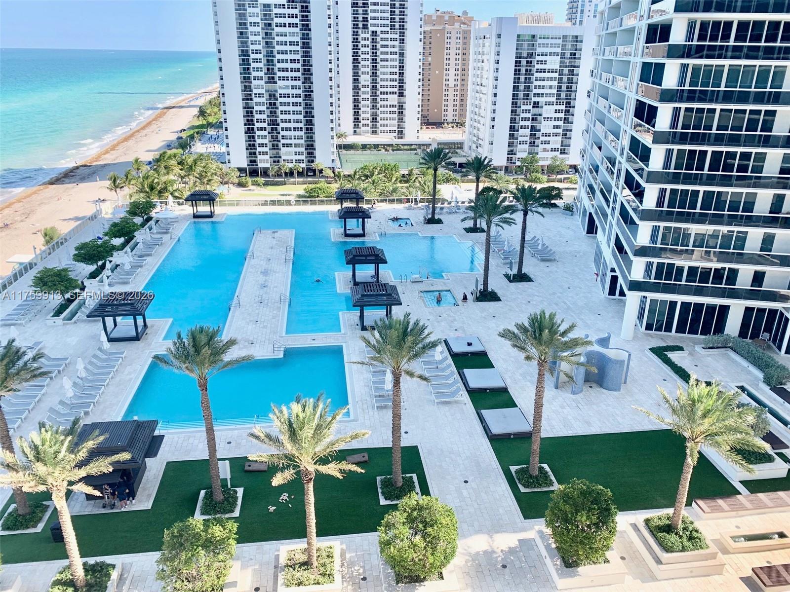 An elevated view of a large outdoor swimming pool area with palm trees and lounge chairs — 1830 S Ocean Dr # 1503, Hallandale Beach FL 33009, HALLANDALE BEACH, FL
