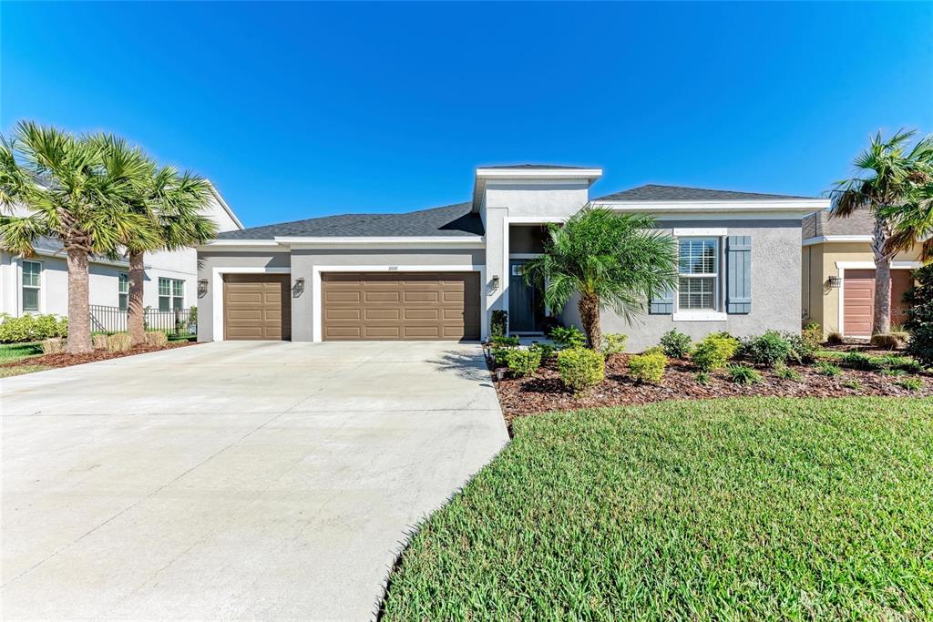 A single-story house with a three-car garage, gray stucco exterior, and palm trees — 10010 BUCKMAN HALLS DRIVE, PALMETTO, FL
