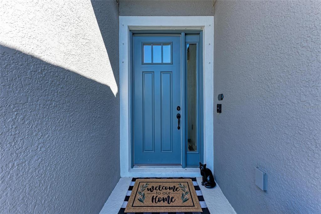 A blue front door with a side window and a welcome mat sits under a porch — 10010 BUCKMAN HALLS DRIVE, PALMETTO, FL