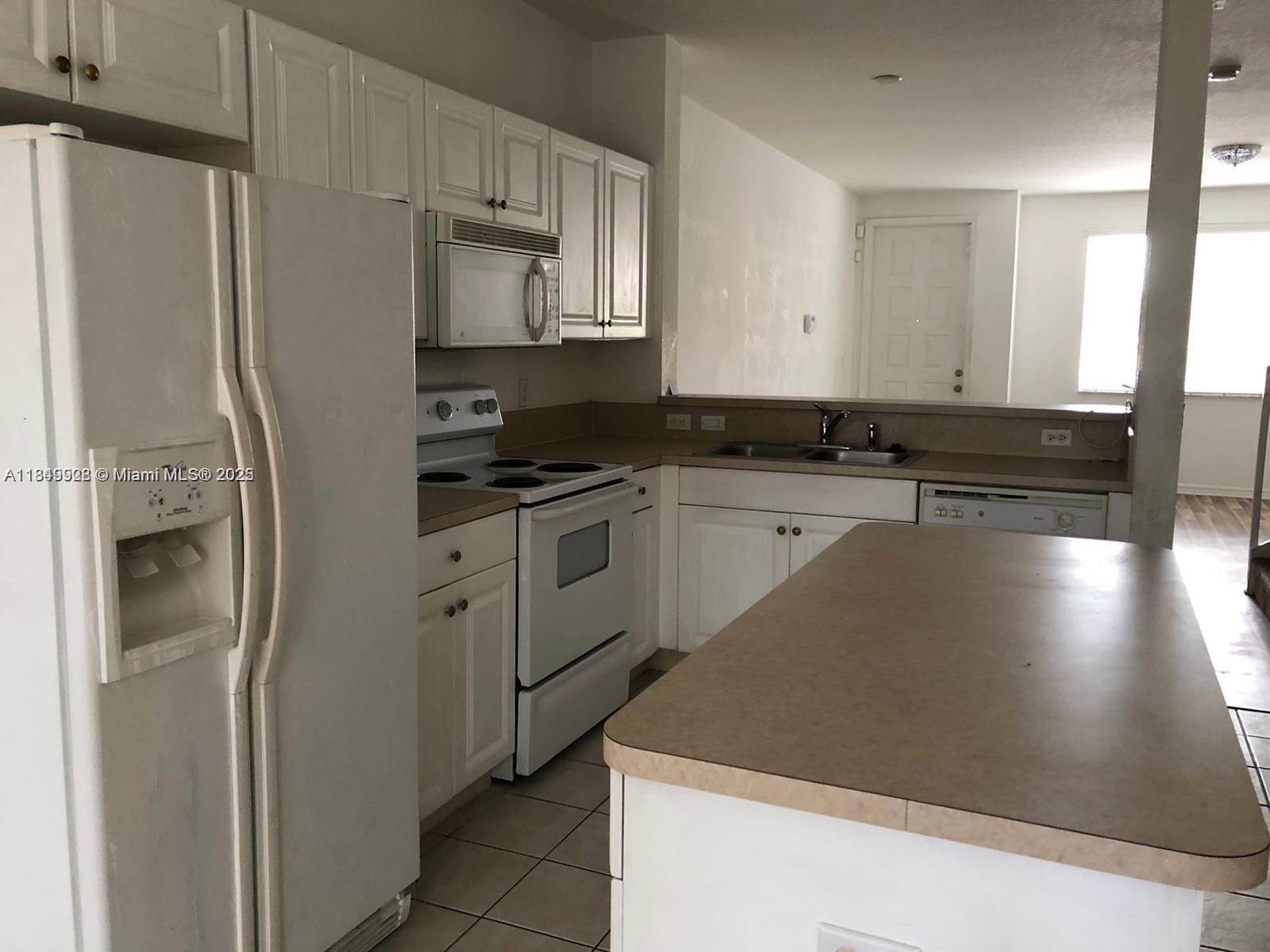 A kitchen features white cabinets, white appliances, a kitchen island, and tan laminate countertops — 1911 Hibiscus Ln, Riviera Beach FL 33404, RIVIERA BEACH, FL