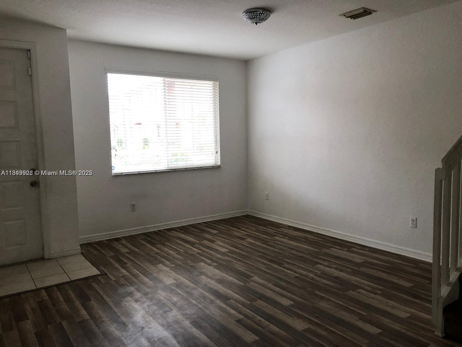 An empty room features white walls, wood-look flooring, a window with blinds, and a staircase — 1911 Hibiscus Ln, Riviera Beach FL 33404, RIVIERA BEACH, FL