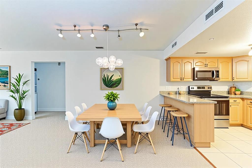 A dining area with a wooden table and white chairs sits next to a kitchen — 3411 CLARK ROAD UNIT 212, SARASOTA, FL