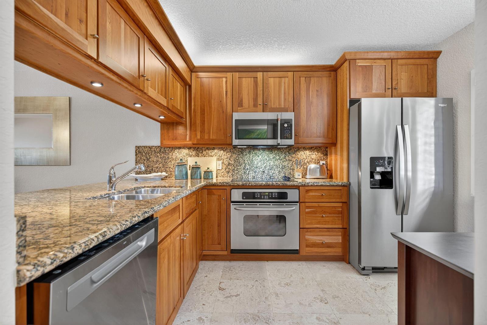 A kitchen features wood cabinets, granite countertops, stainless steel appliances, and light-colored tile flooring — 401 Ocean Bluffs Boulevard # 205, Jupiter FL 33477, JUPITER, FL
