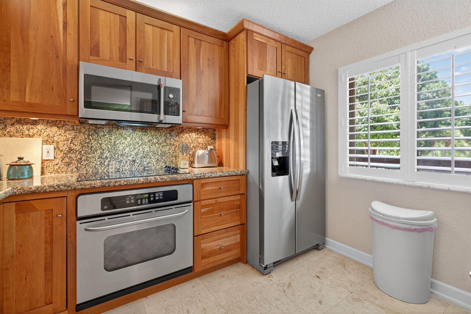 A kitchen features wood cabinets, stainless steel appliances, granite countertops, and a window with shutters — 401 Ocean Bluffs Boulevard # 205, Jupiter FL 33477, JUPITER, FL