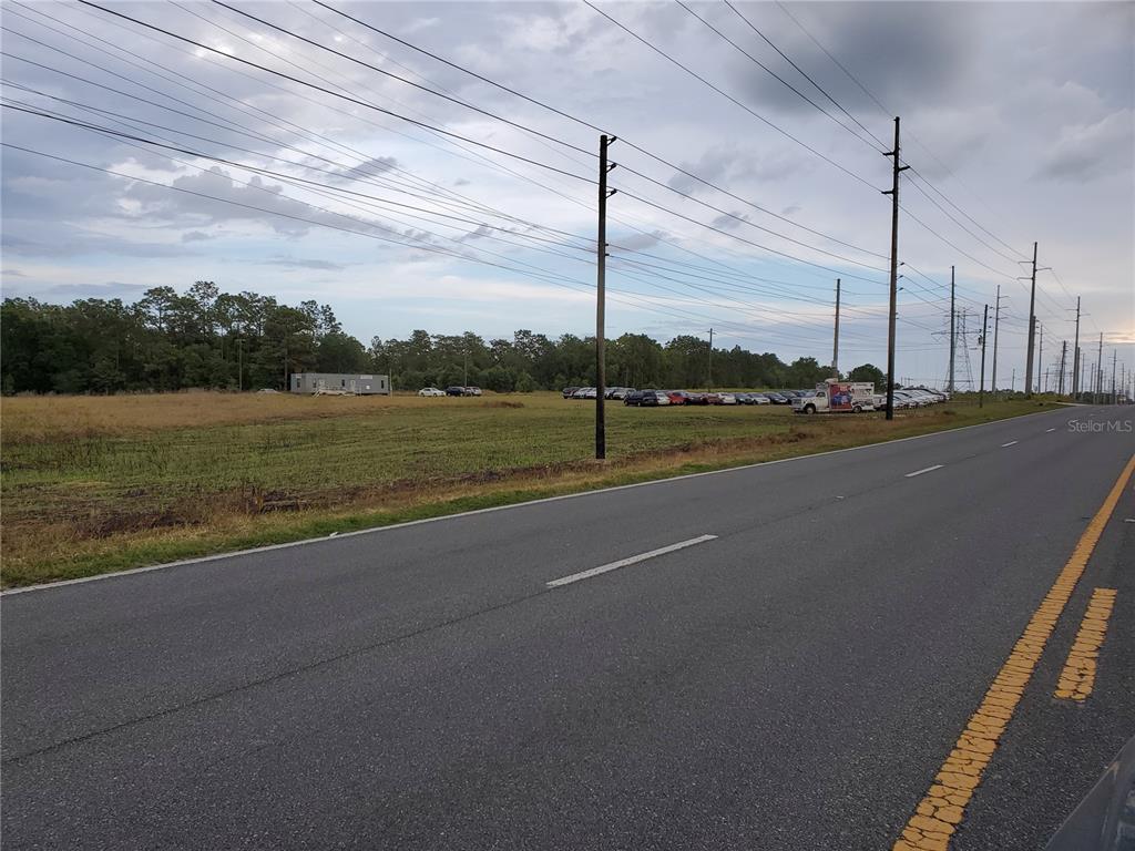 A paved road runs alongside a large, grassy field containing parked cars and a small building — LOT 15 & 16 SE MARICAMP ROAD, OCALA, FL