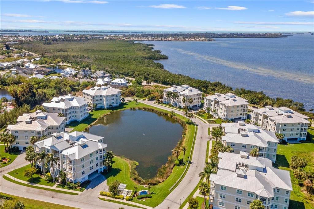 An aerial view shows several multi-story apartment buildings surrounding a central pond near water — 3418 79TH STREET CIRCLE W UNIT 303, BRADENTON, FL