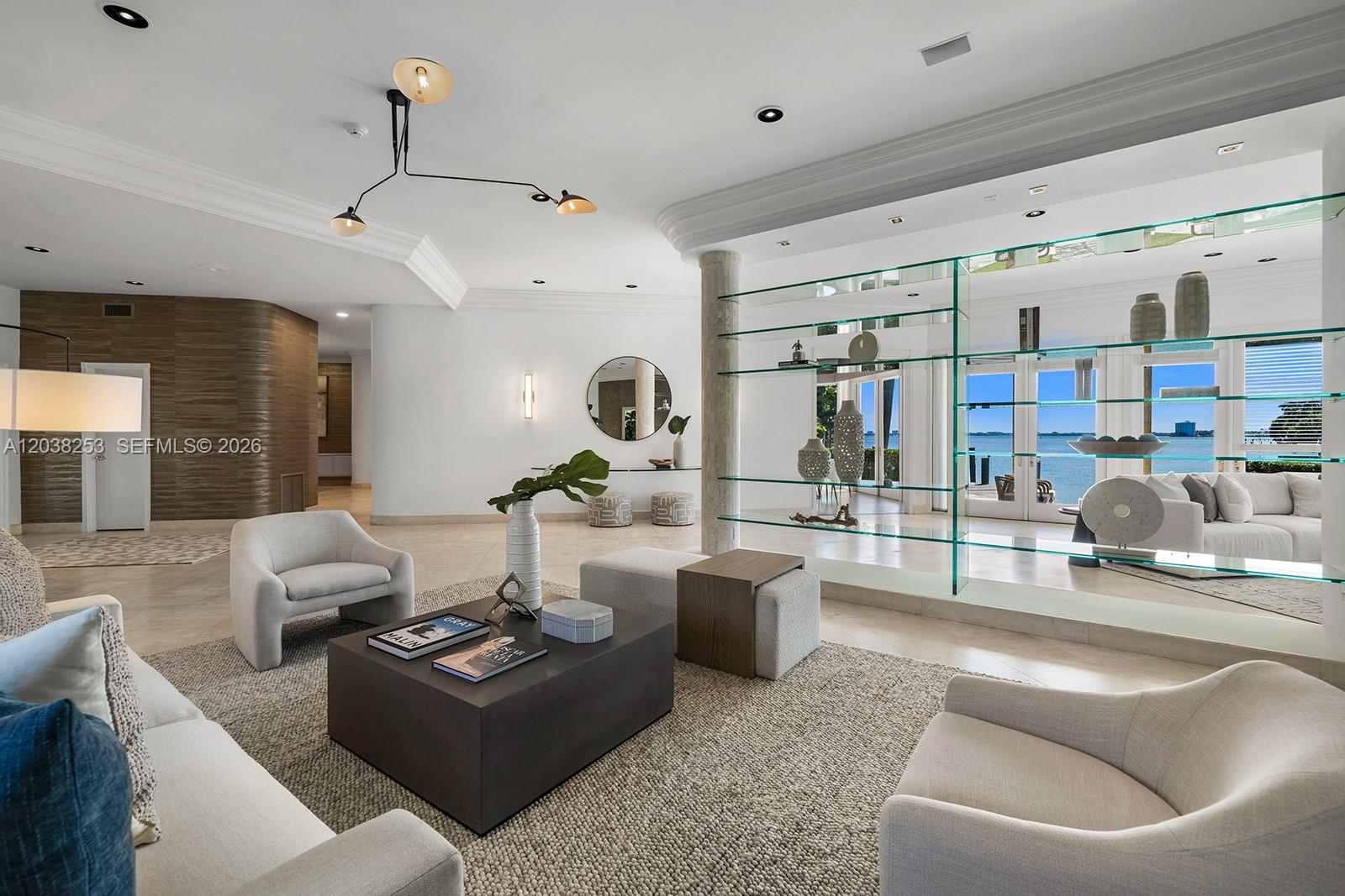 A modern living room features a glass shelving unit, neutral furniture, and a water view — 6500 N Bay Rd, Miami Beach FL 33141, MIAMI BEACH, FL