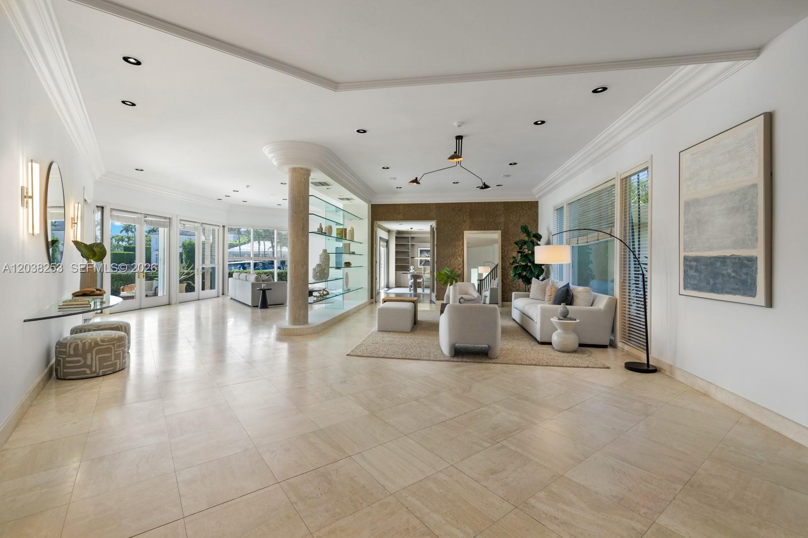 A spacious, open-concept living room features light tile flooring, a central glass shelving unit, and neutral furniture — 6500 N Bay Rd, Miami Beach FL 33141, MIAMI BEACH, FL